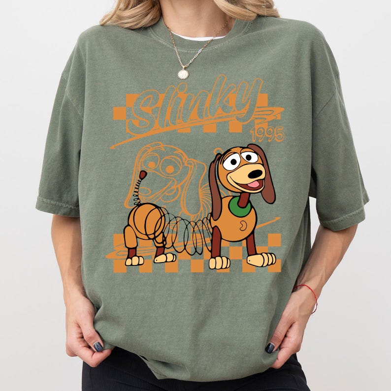 May include: Sage green t-shirt featuring a cartoon image of Slinky Dog with the text "Slinky 1995" in orange. The design includes a checkered orange and white background. The shirt has short sleeves and a crew neck.