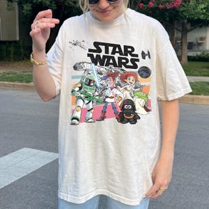 May include: Cream-colored t-shirt featuring a graphic of Star Wars characters in a cartoon style. The design includes the Star Wars logo and images of characters like Darth Vader, Woody, and Buzz Lightyear. The shirt has short sleeves.