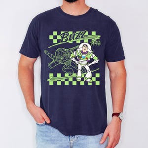 May include: Navy blue t-shirt featuring a graphic of Buzz Lightyear from Toy Story. The design includes the word "Buzz" in green, the year "1995", and a green and black checkered pattern. Buzz Lightyear is depicted in his signature white and green spacesuit.