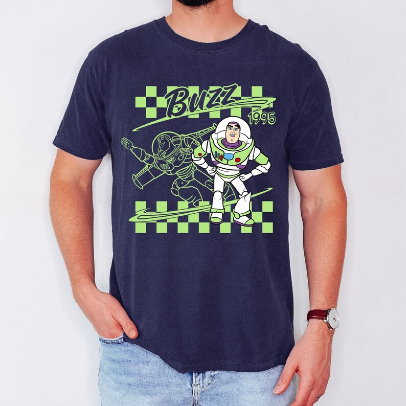 May include: Navy blue t-shirt featuring a graphic of Buzz Lightyear from Toy Story. The design includes the word "Buzz" in green, the year "1995", and a green and black checkered pattern. Buzz Lightyear is depicted in his signature white and green spacesuit.