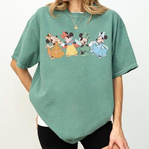 Puede incluir: Camiseta verde con un estampado de cuatro personajes de Minnie Mouse con vestidos coloridos. Los personajes de dibujos animados son en tonos amarillos, rojos, verdes y azules. La camiseta tiene mangas cortas y un corte holgado.