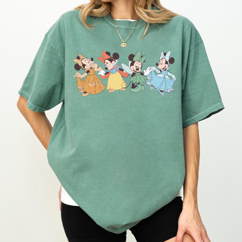 Puede incluir: Camiseta verde con un estampado de cuatro personajes de Minnie Mouse con vestidos coloridos. Los personajes de dibujos animados son en tonos amarillos, rojos, verdes y azules. La camiseta tiene mangas cortas y un corte holgado.