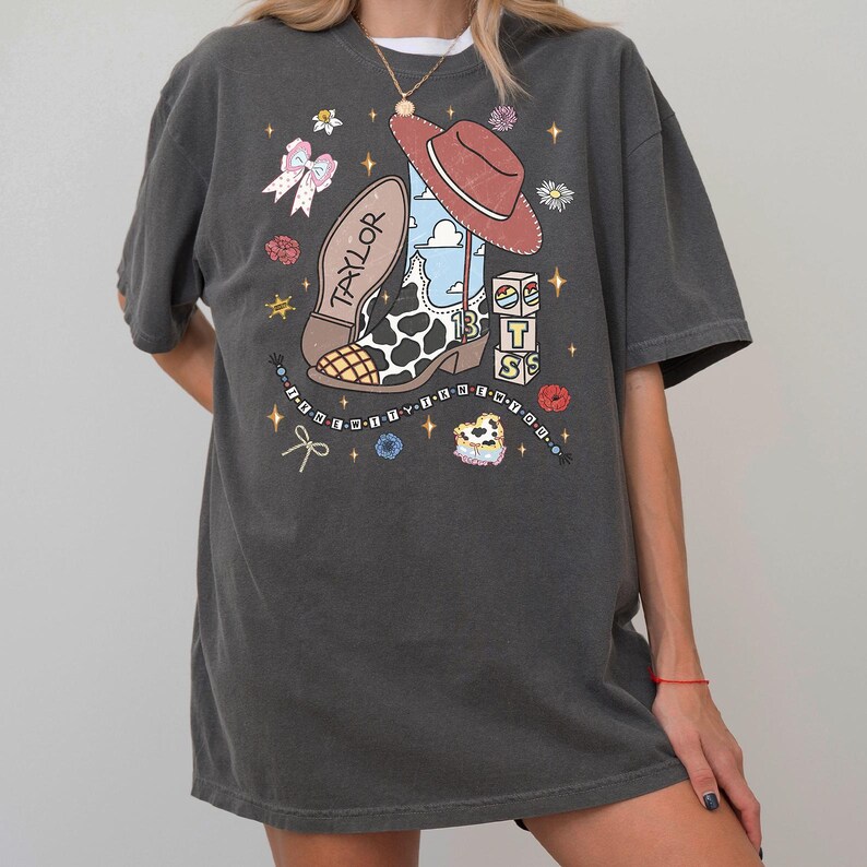 May include: A charcoal gray t-shirt featuring a graphic print of a cow-print cowboy boot, a brown hat, and blocks with the letters T, S, and the number 13. The design includes the name Taylor, a beaded string, bows, and small floral accents.