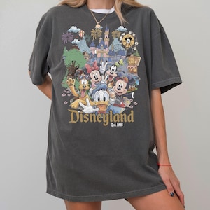 May include: A charcoal gray short-sleeve t-shirt featuring a colorful graphic print of various cartoon characters in front of a castle. The text "Disneyland Est. 1955" is printed in gold lettering below the characters, which include a mouse, duck, and dog.