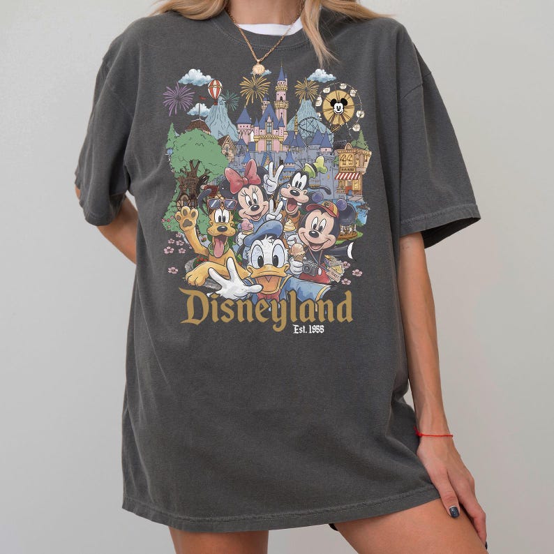 May include: A charcoal gray short-sleeve t-shirt featuring a colorful graphic print of various cartoon characters in front of a castle. The text "Disneyland Est. 1955" is printed in gold lettering below the characters, which include a mouse, duck, and dog.