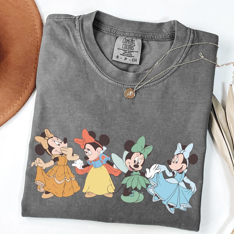 Puede incluir: Una camiseta gris Comfort Colors con un gr&aacute;fico de cuatro personajes de Minnie Mouse con vestidos coloridos. Un collar dorado con un colgante descansa sobre la camiseta. La camiseta est&aacute; doblada sobre una superficie blanca.