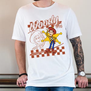 May include: White t-shirt featuring a cartoon graphic of Woody from Toy Story. The design includes a sheriff's badge, the word "Woody," and the year "1995" in brown. The shirt has short sleeves and a crew neck.