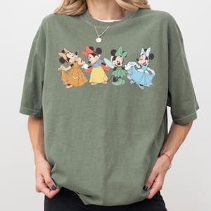 Puede incluir: Camiseta verde oliva con un gr&aacute;fico de cuatro personajes de Minnie Mouse con vestidos. Los personajes de dibujos animados visten vestidos amarillos, rojos, verdes y azules. La camiseta tiene mangas cortas y cuello redondo.