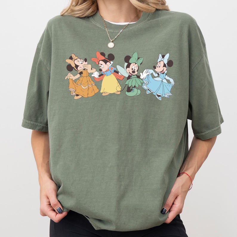 Puede incluir: Camiseta verde oliva con un gr&aacute;fico de cuatro personajes de Minnie Mouse con vestidos. Los personajes de dibujos animados visten vestidos amarillos, rojos, verdes y azules. La camiseta tiene mangas cortas y cuello redondo.