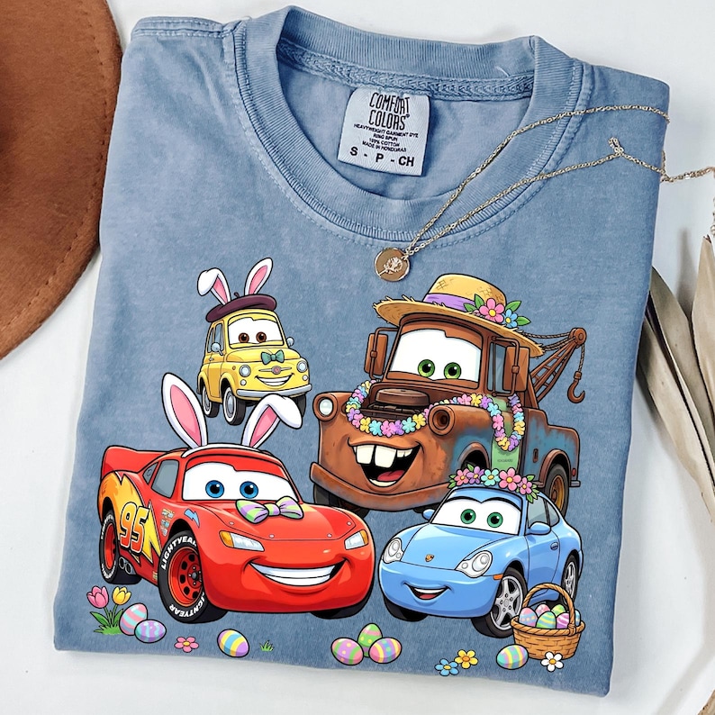 Puede incluir: Una camiseta azul Comfort Colors con un gr&aacute;fico de coches animados con orejas de conejo y huevos de Pascua. Los coches son rojos, amarillos, marrones y azules. Un collar dorado est&aacute; sobre la camiseta.