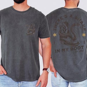 May include: A charcoal gray t-shirt featuring a small graphic on the front left chest of a cowboy boot with a snake. The back displays a large graphic of a cowboy boot with a snake, the text "THERE'S A SNAKE IN MY BOOT", "EST. 1995", and "Yee-Haw".