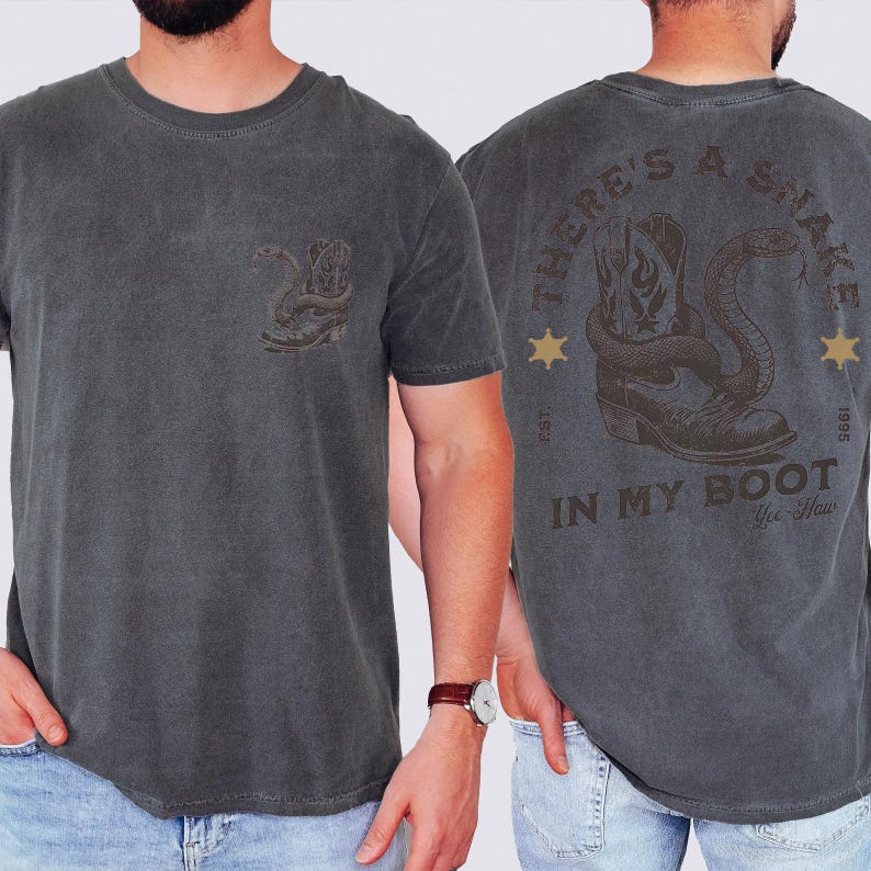 May include: A charcoal gray t-shirt featuring a small graphic on the front left chest of a cowboy boot with a snake. The back displays a large graphic of a cowboy boot with a snake, the text "THERE'S A SNAKE IN MY BOOT", "EST. 1995", and "Yee-Haw".