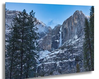 Könnte beinhalten: Landschaftsfotografie eines Gebirges mit einem Wasserfall. Die Berge sind teilweise schneebedeckt, und immergrüne Bäume rahmen die Szene ein. Der Himmel ist ein klares Blau.