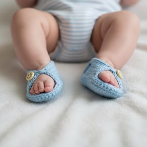May include: A pair of light blue crochet baby sandals with a button featuring a smiling face on each sandal. The sandals are placed on a white surface, with the baby's feet and legs visible.