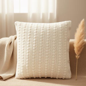 Peut inclure: Un coussin décoratif carré blanc cassé avec un motif tricoté texturé. Le coussin est posé sur un canapé beige, avec une couverture assortie drapée sur le bras. Une plante séchée de couleur beige ajoute une touche naturelle.