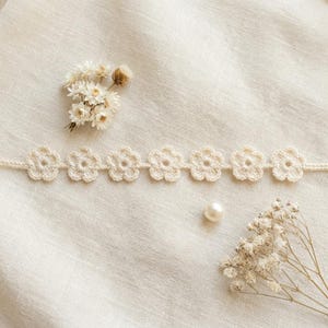 May include: A cream-colored, crocheted floral choker necklace. The necklace features seven flower-shaped designs. The image also includes dried flowers and a pearl on a white linen background.