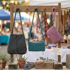Puede incluir: Tres bolsos de crochet hechos a mano y una pequeña bolsa se exhiben en un estante de madera. Los bolsos son en tonos grises, verde azulado y malva. El letrero dice "HANDMADE CROCHET PATTERN - Etsy". Los bolsos tienen correas largas y una puntada texturizada.