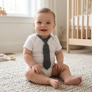 May include: A baby wearing a white onesie and a grey crocheted tie smiles, sitting on a textured rug. A wooden cot and toys are in the background, suggesting a nursery. The room has a neutral colour palette.