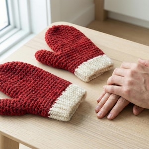 May include: A pair of red knitted mittens with off-white cuffs, resting on a light wooden surface. The mittens are handmade, with a textured appearance. The image is well-lit, showcasing the details of the yarn and stitch work.