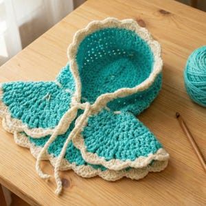 May include: A handmade turquoise crochet bonnet with a scalloped edge and cream trim, tied with cream yarn. A ball of turquoise yarn and a wooden crochet hook sit on a wooden table, suggesting a crafting theme.