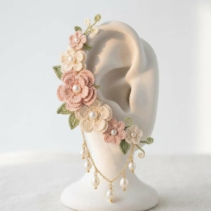 May include: An ear cuff featuring crocheted flowers in shades of dusty pink and cream, accented with small green crocheted leaves. The design includes white pearl centers and a delicate gold-toned chain with dangling white pearl beads.