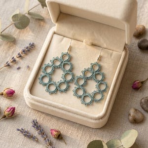 Puede incluir: Un par de pendientes en forma de lágrima de color verde salvia, elaborados con intrincado encaje de frivolité. Cada pendiente presenta una serie de bucles circulares interconectados con delicados bordes de picot, suspendidos de ganchos de metal plateado.