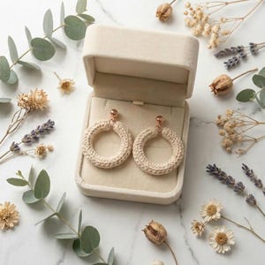 May include: A pair of circular earrings featuring a beige crocheted yarn covering over a hoop frame, attached to a small rose gold-toned metal ball stud. The earrings are presented inside an open, rectangular, cream-colored velvet jewelry box.