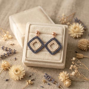 May include: A pair of dangle earrings featuring two nested square frames in cream and dark gray thread, suspended from a small rose gold-toned metal ball stud. The earrings are displayed on a square, beige velvet jewelry box insert.