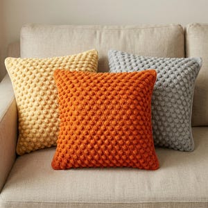 Pode incluir: Quatro almofadas decorativas quadradas em tons de laranja, amarelo e cinza. As almofadas têm um padrão de pontos em relevo e estão dispostas em um sofá bege. As almofadas adicionam um toque aconchegante e colorido à decoração da casa.