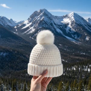 May include: A cream-colored knit beanie with a fluffy white pom-pom. The hat has a ribbed brim and a textured body. The background features a mountain range with snow-covered peaks and a clear blue sky.