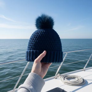 May include: A navy blue knit beanie with a matching faux fur pom-pom. The hat is held up against a backdrop of the ocean and a clear blue sky. The ribbed brim and textured body of the hat are clearly visible.