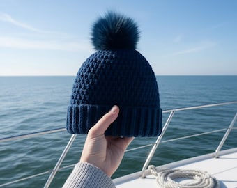 May include: A navy blue knit beanie with a matching faux fur pom-pom. The hat is held up against a backdrop of the ocean and a clear blue sky. The ribbed brim and textured body of the hat are clearly visible.