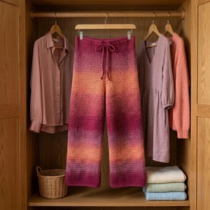 May include: A pair of wide-leg, crochet pants with a gradient color scheme, transitioning from orange to deep pink. The pants hang on a wooden hanger in a closet, alongside other clothing items. A small woven basket sits on a shelf below.