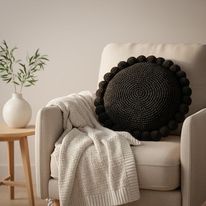 May include: A dark brown, round, knitted pillow with a pom-pom border rests on a cream-colored armchair. A cream-colored knitted throw blanket is draped over the chair. A white vase with green stems sits on a small wooden table.