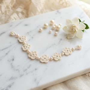May include: A cream-coloured crochet bracelet with flower-shaped designs and small pearl accents. The bracelet is displayed on a marble surface, with loose pearls and a white gardenia flower. The bracelet is a delicate piece of jewellery.