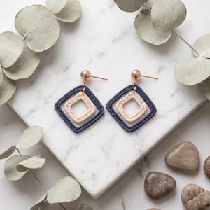 May include: A pair of drop earrings featuring two nested square frames made of crocheted thread. The outer square is dark navy blue, while the inner square is cream. The earrings are attached to rose gold-toned metal ball stud posts.