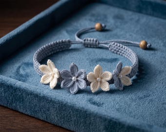 May include: A woven bracelet featuring four crocheted flower motifs in alternating shades of cream and light blue. Each flower is centered with a small white bead. The band is made of braided cord with an adjustable sliding knot and two wooden beads.