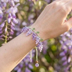 May include: A handmade crochet bracelet featuring a wisteria flower design. The bracelet is primarily lavender and green, with delicate floral details. The bracelet is tied with a light green cord and a small wooden bead.