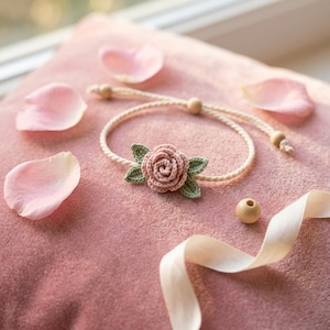 May include: A handmade bracelet featuring a crocheted pink rose with green leaves. The bracelet is made of a cream-colored cord and wooden beads. The bracelet is displayed on a pink surface with scattered rose petals and a cream-colored ribbon.