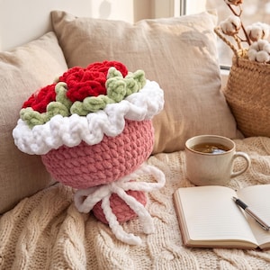 Puede incluir: Un ramo de crochet hecho a mano con rosas rojas, hojas verdes y ribete blanco. El jarrón rosa está atado con un lazo blanco. El ramo está sobre una manta de punto color crema, junto a una taza de té y un cuaderno.