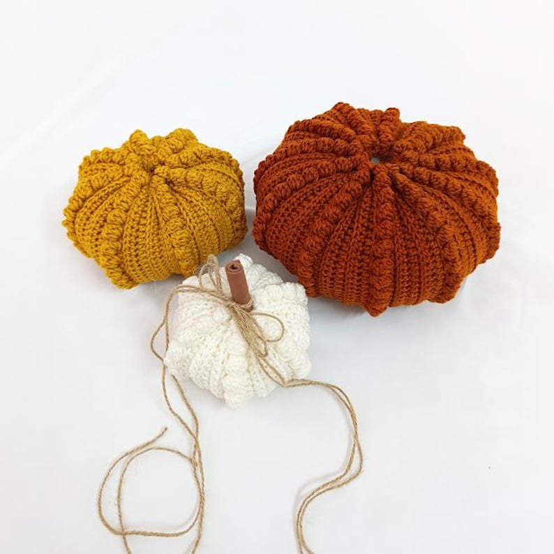 Puede incluir: Tres calabazas de ganchillo en tonos amarillo, naranja y blanco. Las calabazas tienen una textura acanalada y est&aacute;n dispuestas sobre una superficie blanca. La calabaza blanca tiene un tallo de canela y cuerda.