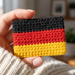 May include: A tablet displaying a digital crochet pattern grid featuring the black, red, and gold colours of the German flag. The screen shows rows of stitch symbols arranged in a rectangular block, with a finger touching the screen surface.
