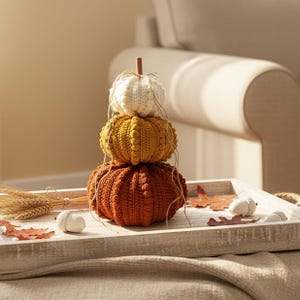 Puede incluir: Una composici&oacute;n oto&ntilde;al con tres calabazas de ganchillo apiladas, crema, dorada y naranja. Se presentan en una bandeja de madera con hojas y bellotas, creando un ambiente de temporada. Un peque&ntilde;o tallo de madera y cuerda completan el conjunto.