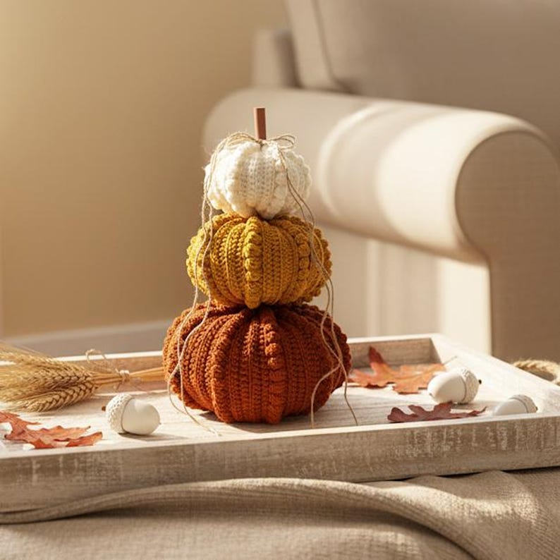 Puede incluir: Una composici&oacute;n oto&ntilde;al con tres calabazas de ganchillo apiladas, crema, dorada y naranja. Se presentan en una bandeja de madera con hojas y bellotas, creando un ambiente de temporada. Un peque&ntilde;o tallo de madera y cuerda completan el conjunto.