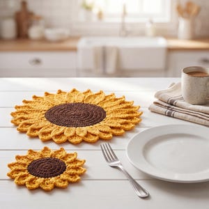 May include: A crocheted sunflower placemat and coaster set. The placemat is a large, round design with yellow petals and a brown center. The coaster mirrors the design. A white plate, fork, and mug are also on the table.