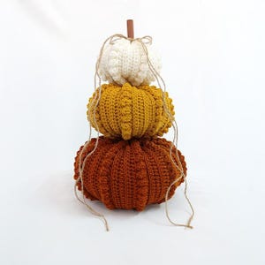 Puede incluir: Una pila decorativa de tres calabazas de ganchillo en crema, amarillo mostaza y naranja &oacute;xido. Las calabazas est&aacute;n dispuestas verticalmente, con un tallo de madera y detalles de cuerda. Las calabazas est&aacute;n hechas a mano y tienen una superficie texturizada.