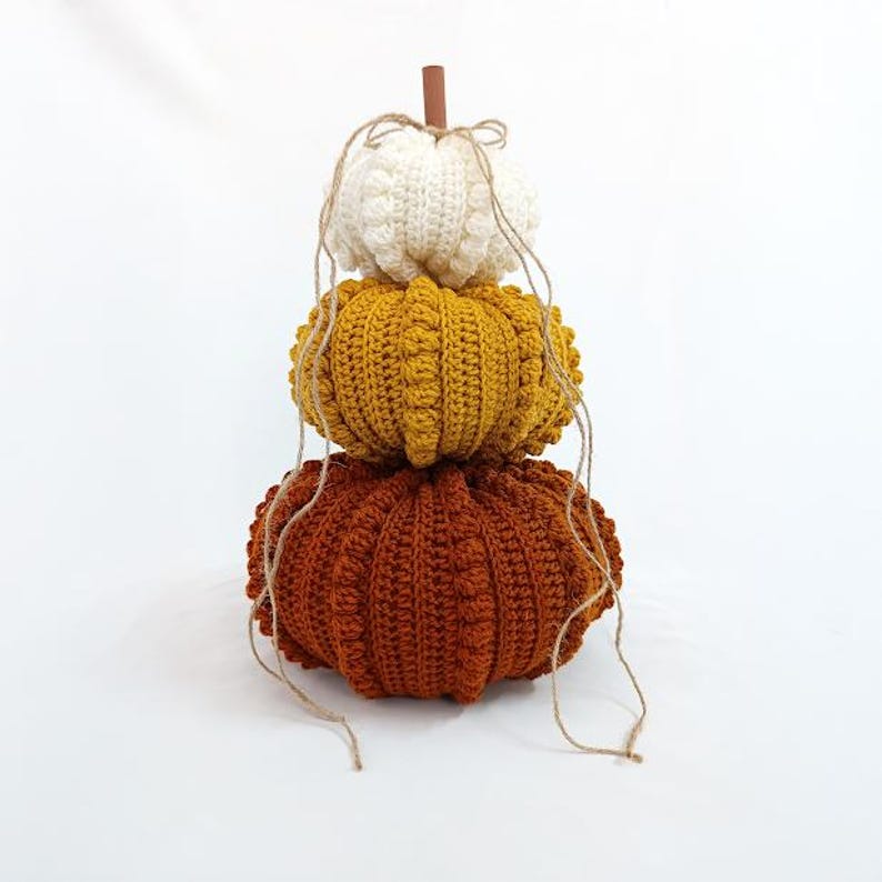 Puede incluir: Una pila decorativa de tres calabazas de ganchillo en crema, amarillo mostaza y naranja &oacute;xido. Las calabazas est&aacute;n dispuestas verticalmente, con un tallo de madera y detalles de cuerda. Las calabazas est&aacute;n hechas a mano y tienen una superficie texturizada.