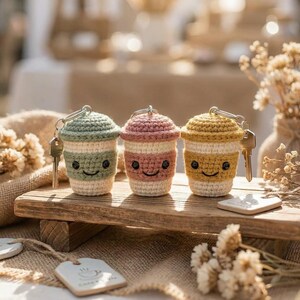 May include: Three crocheted coffee cup keychains in pastel colors: green, pink, and yellow. Each keychain has a smiling face and a key attached. They are displayed on a wooden surface.