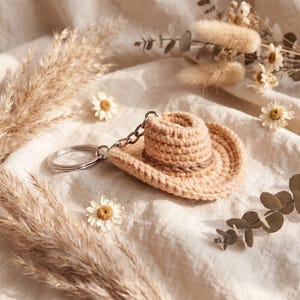 May include: A crocheted keychain shaped like a miniature cowboy hat in a light tan color, with a darker brown band. The hat is attached to a silver-colored chain and keyring. The background features dried flowers and a soft, neutral-toned fabric.