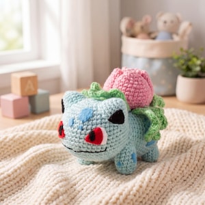 May include: A crocheted Bulbasaur plush toy in light blue, with a pink flower on its back. The toy has black eyes, red accents, and blue spots. It is sitting on a cream-colored knitted blanket.